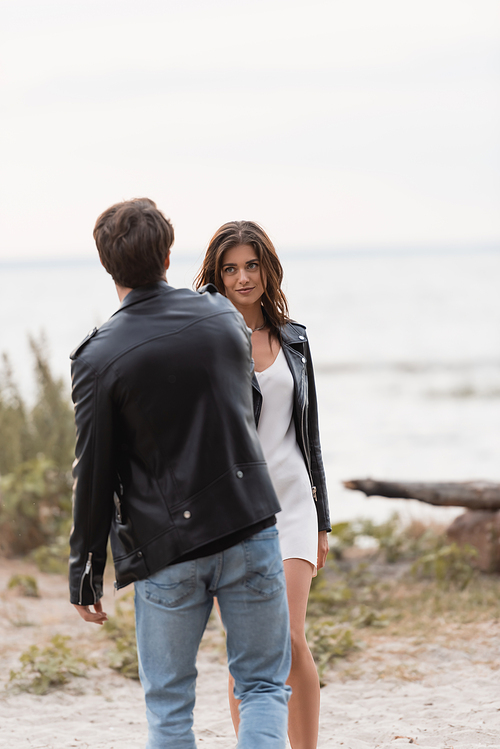 Selective focus of young woman in dress and leather jacket looking at boyfriend on beach