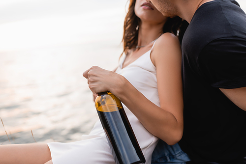 Cropped view of man kissing girlfriend in dress with bottle of wine near sea