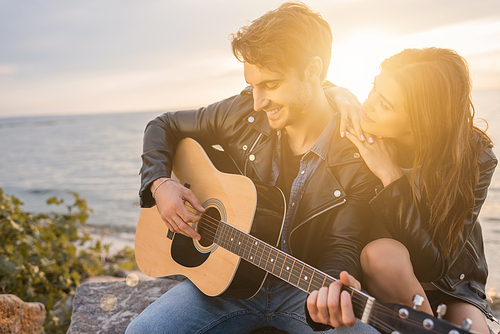 Young man in leather jacket playing acoustic guitar near girlfriend on beach at sunset