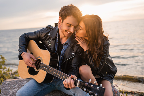 Young woman kissing boyfriend playing acoustic guitar on seaside during sunset