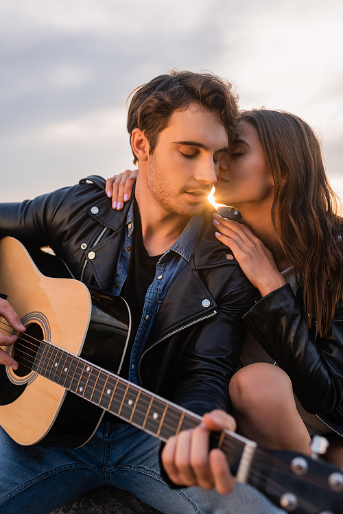 Selective focus of woman kissing boyfriend with acoustic guitar at sunset