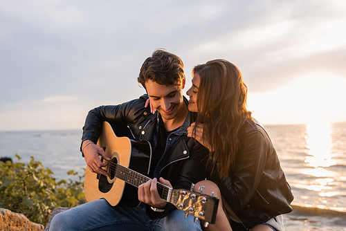 Selective focus of woman in leather jacket embracing boyfriend playing acoustic guitar during sunset on beach