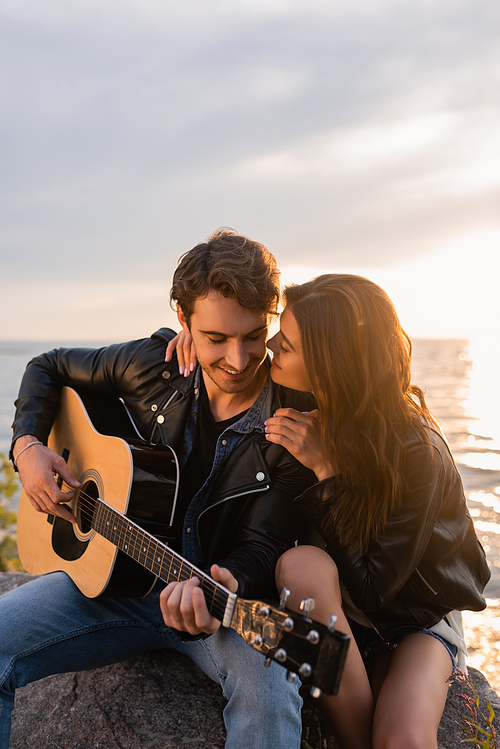 Selective focus of woman hugging boyfriend playing acoustic guitar near sea during sunset