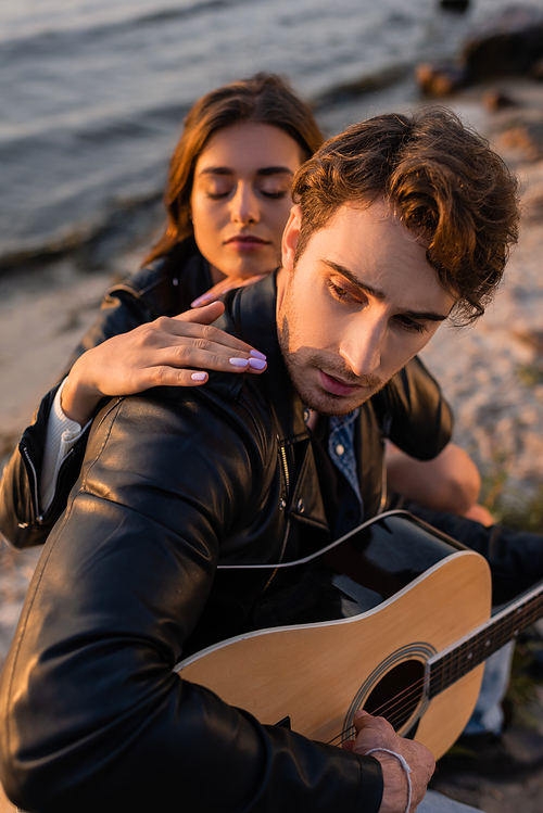 Selective focus of man playing acoustic guitar near woman on beach near sea at sunset