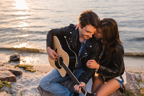Brunette woman hugging boyfriend in leather jacket with acoustic guitar on seaside at evening