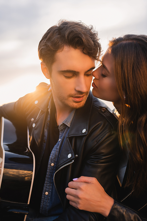Brunette woman kissing man in leather jacket with acoustic guitar at sunset