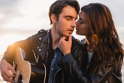 Brunette woman kissing boyfriend in leather jacket with acoustic guitar during sunset