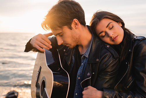 Young man holding acoustic guitar near woman on beach at sunset