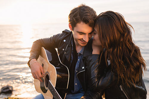 Woman embracing boyfriend playing acoustic guitar near sea on beach at evening