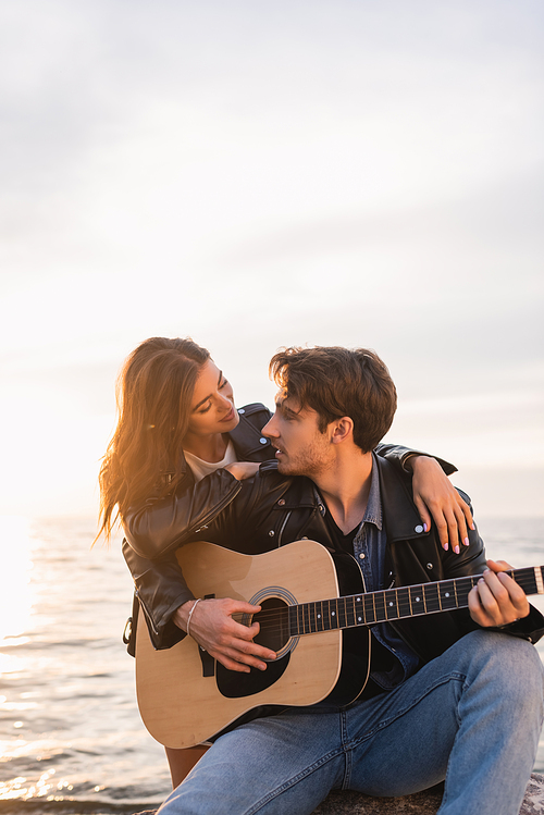 Young woman in leather jacket embracing boyfriend playing acoustic guitar near sea at sunset