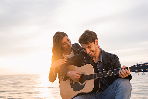 Woman hugging boyfriend playing acoustic guitar near sea and sunset at background