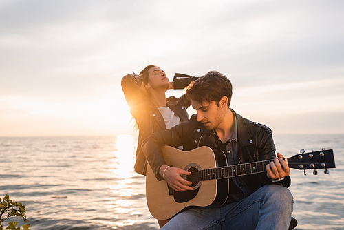 Selective focus of man in leather jacket playing acoustic guitar near girlfriend during sunset near sea