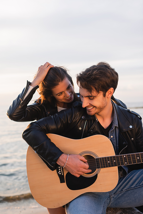 Burnette woman touching hair and looking at boyfriend playing acoustic guitar on beach at evening