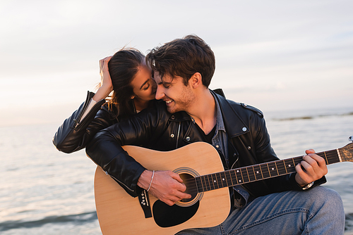 Young woman kissing boyfriend playing acoustic guitar near sea at sunset