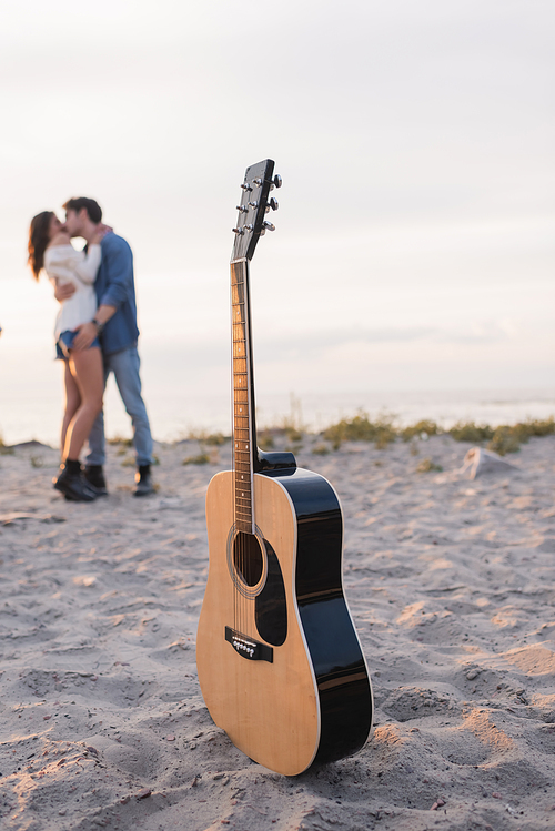 Selective focus of acoustic guitar on sand and young couple kissing on beach at sunset
