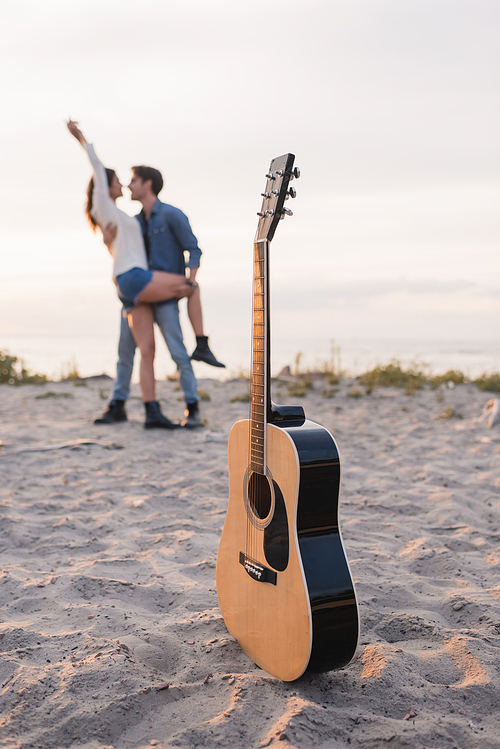 Selective focus of acoustic guitar on sand near young couple embracing on beach at sunset