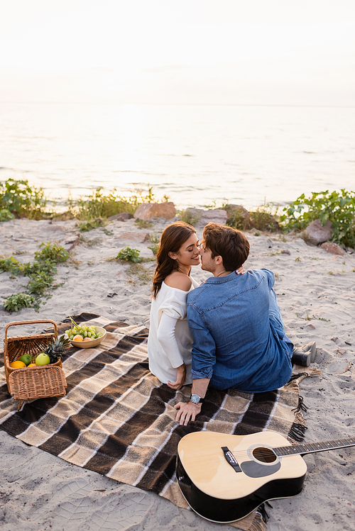Young couple kissing near acoustic guitar and wicker basket during picnic on beach