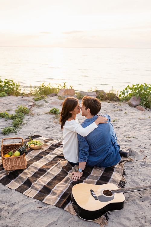 Selective focus of couple kissing near acoustic guitar during picnic on sea shore at sunset
