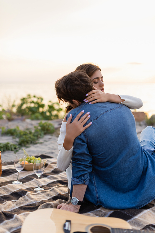 Selective focus of woman embracing boyfriend near acoustic guitar and wine glasses on beach