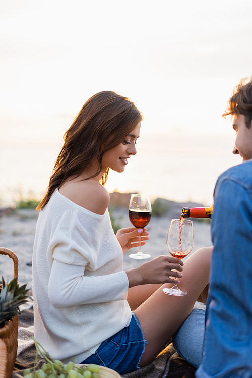 Selective focus of man pouring wine near girlfriend with glasses on beach at sunset