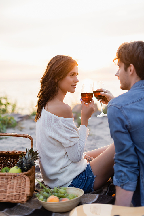 Selective focus of couple clinking with wine during picnic on beach at sunset