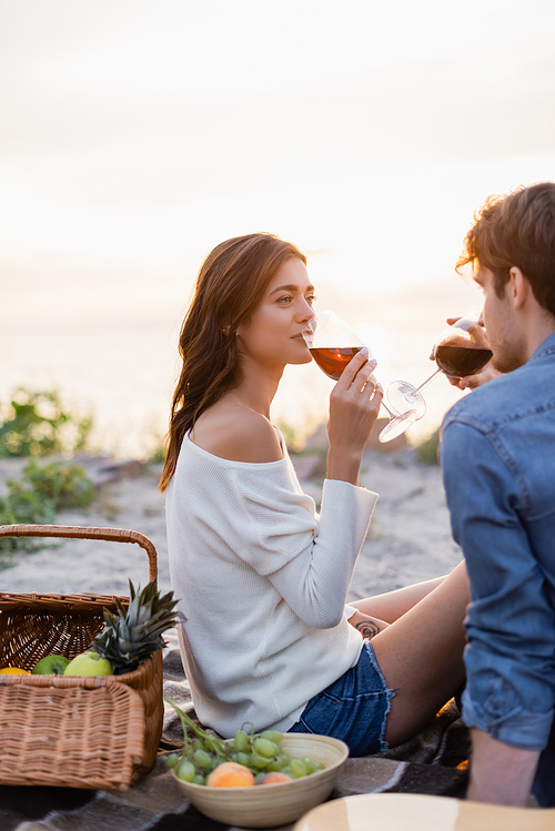 Selective focus of woman drinking wine near boyfriend during picnic on beach at sunset