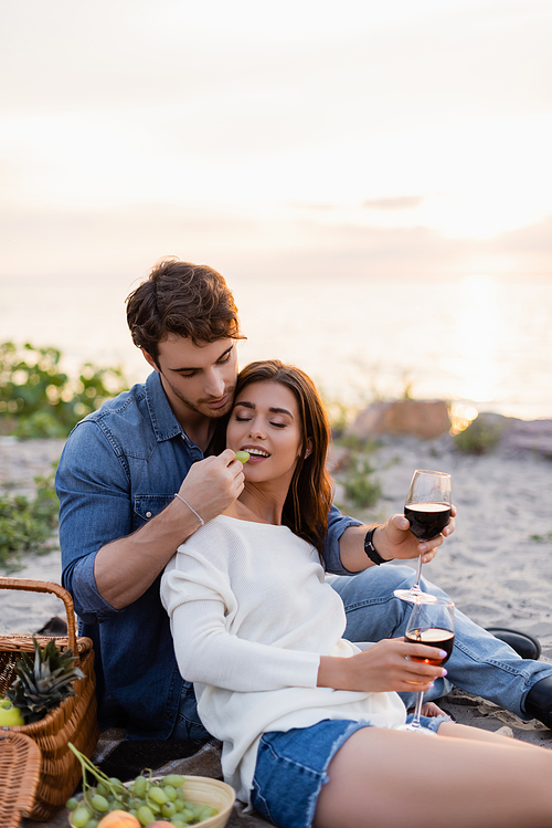 Selective focus of man feeding girlfriend with grape while holding glass of wine on beach