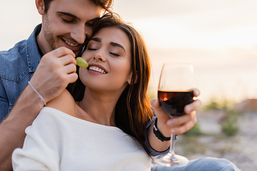 Selective focus of man feeding girlfriend with grape while holding glass of wine on beach