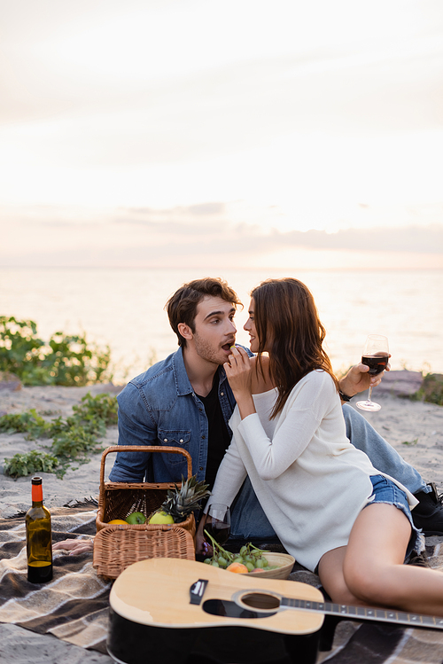 Selective focus of woman feeding boyfriend with grape near wine, fruits and acoustic guitar on blanket on beach