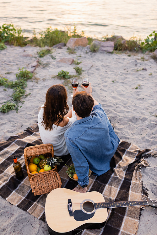 Overhead view of couple holding glasses of wine during picnic near acoustic guitar during sunset