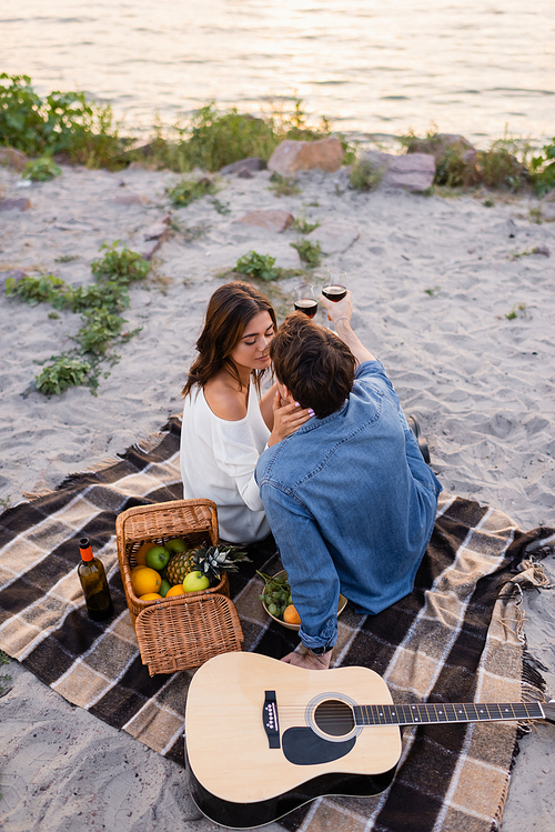 Overhead view of couple kissing while holding glasses of wine near acoustic guitar on beach