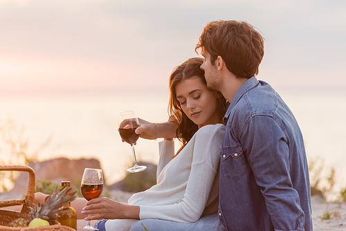 Selective focus of man hugging girlfriend with glass of wine during picnic on beach