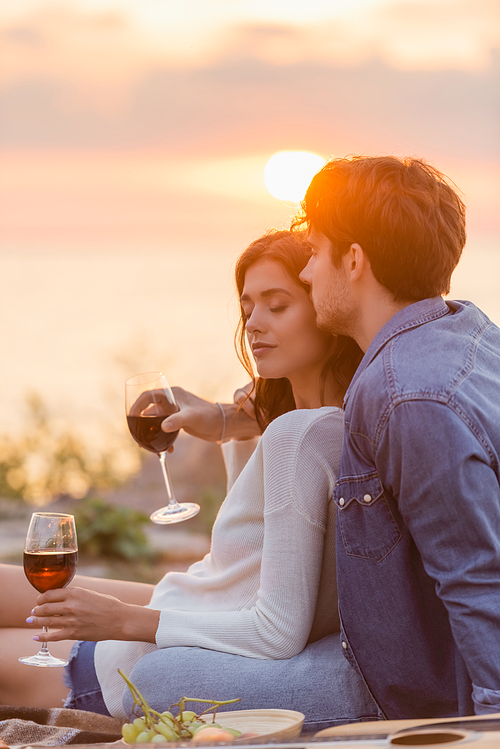 Selective focus of man embracing girlfriend with glass of wine near acoustic guitar on beach