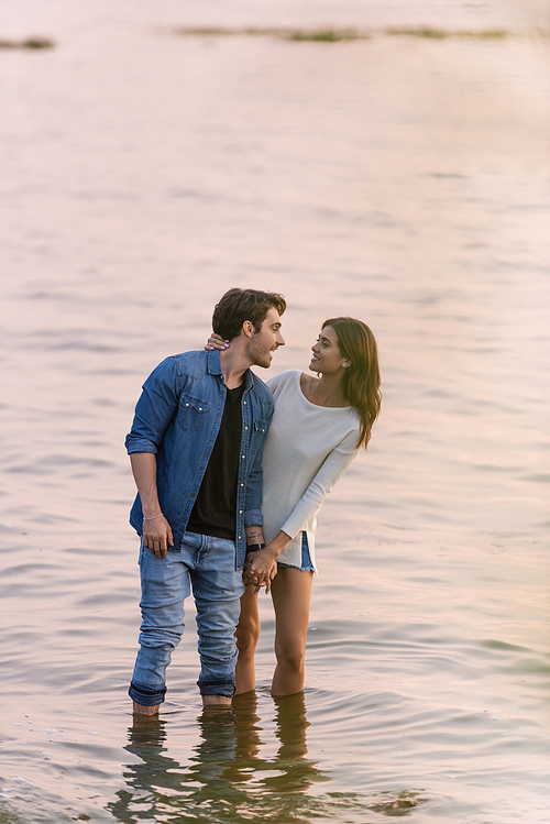Brunette woman touching boyfriend while standing in sea water at sunset