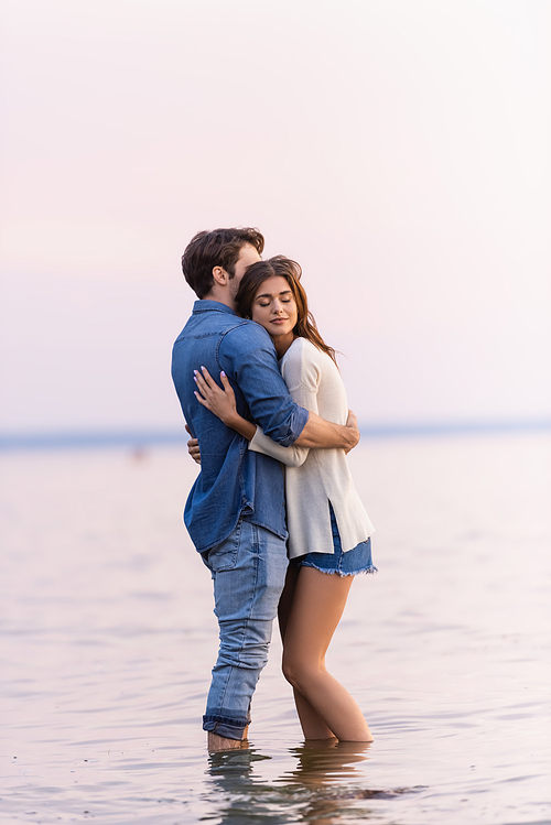 Side view of brunette woman with closed eyes hugging boyfriend while standing in sea