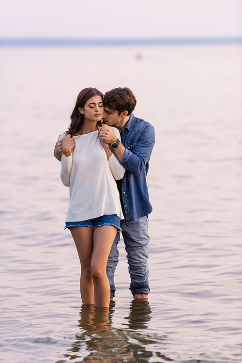 Man hugging brunette girlfriend with closed eyes while standing in sea at sunset