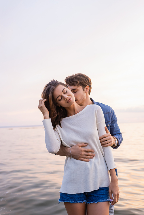 Young man kissing neck and hugging brunette woman near sea