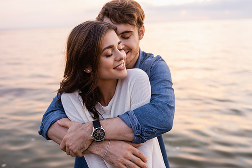 Young man hugging brunette girlfriend with closed eyes beside sea at sunset