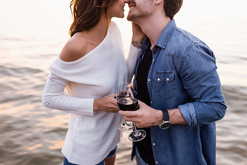 Cropped view of young couple kissing while holding glasses of wine near sea