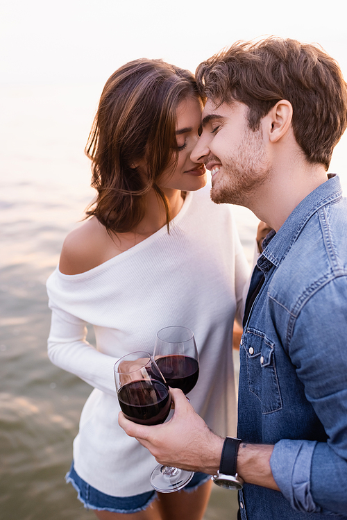 Young man with glass of wine standing near girlfriend and sea at sunset