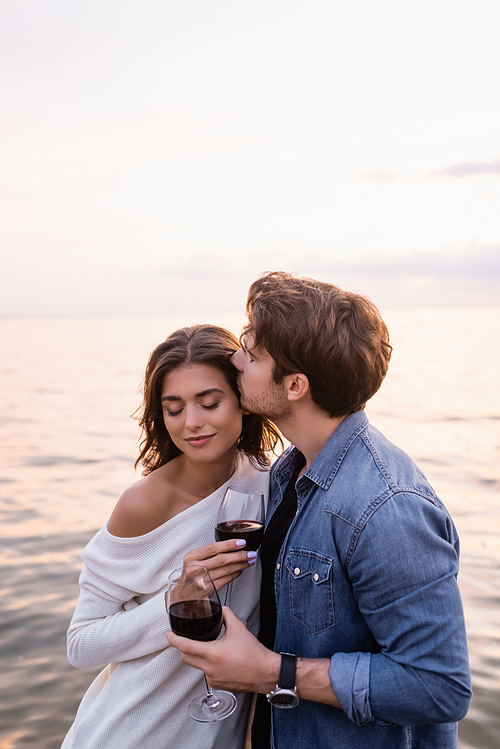 Young man kissing girlfriend with glass of wine near sea at evening