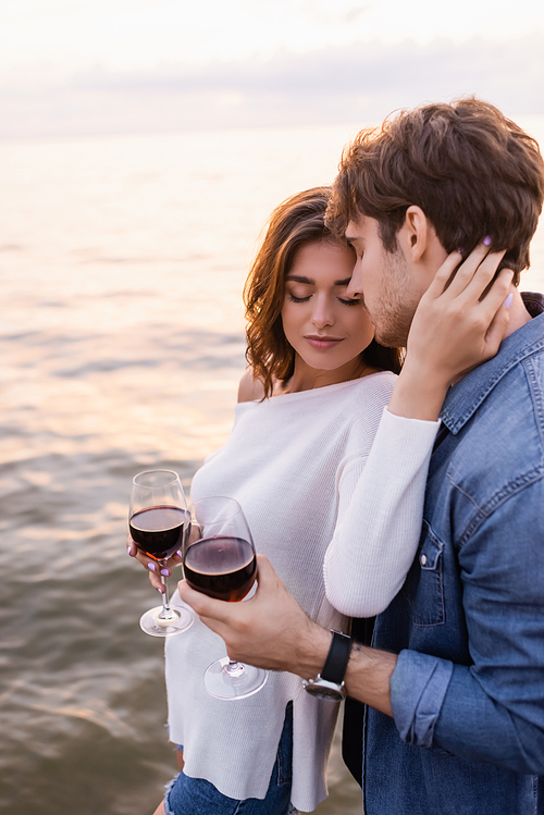 Brunette woman holding glass of red wine and touching neck of boyfriend near sea