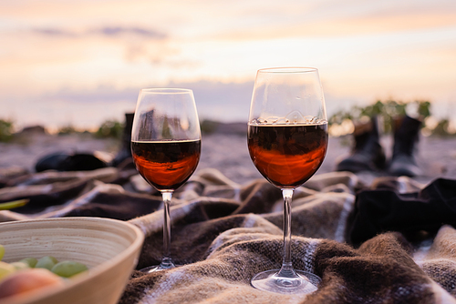 Selective focus of glasses of wine near fruits in bowl on plaid on beach