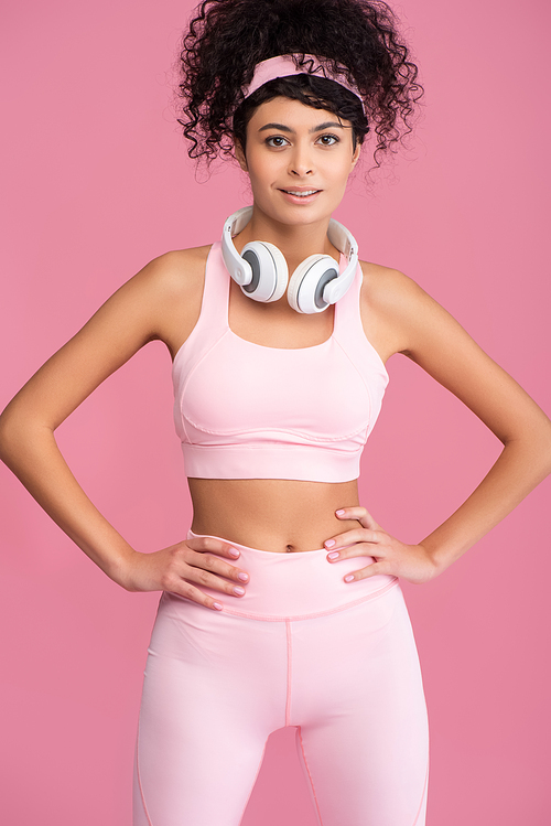 curly young woman in wireless headphones standing with hands on hips isolated on pink