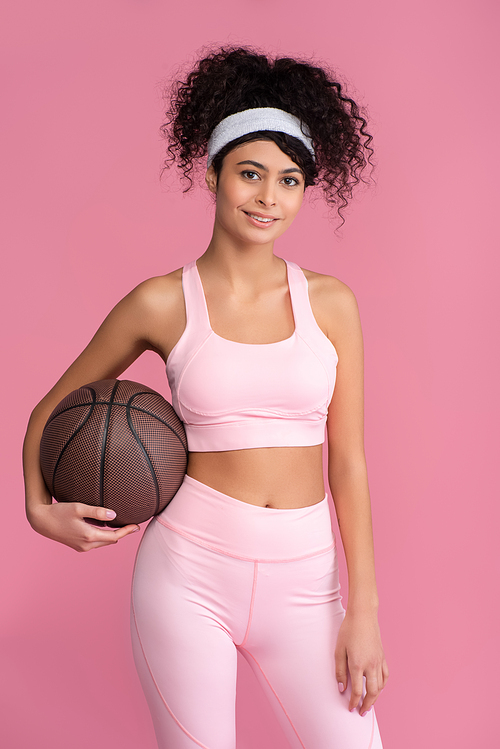 cheerful young woman holding basketball isolated on pink