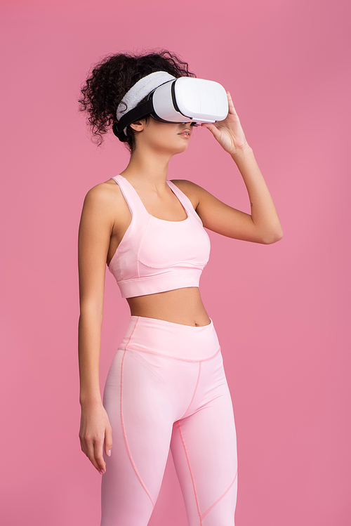 curly woman touching virtual reality headset isolated on pink