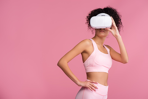 curly woman in virtual reality headset standing with hand on hip isolated on pink