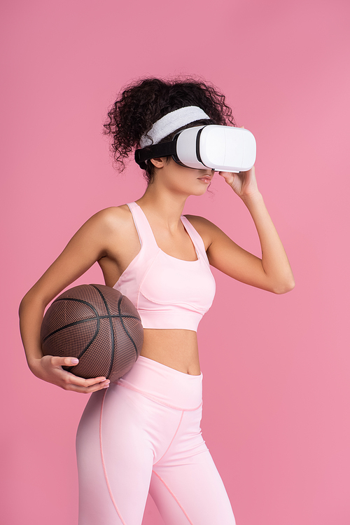 curly woman in virtual reality headset holding basketball isolated on pink