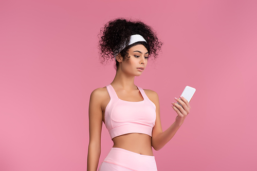 curly young sportswoman using smartphone isolated on pink