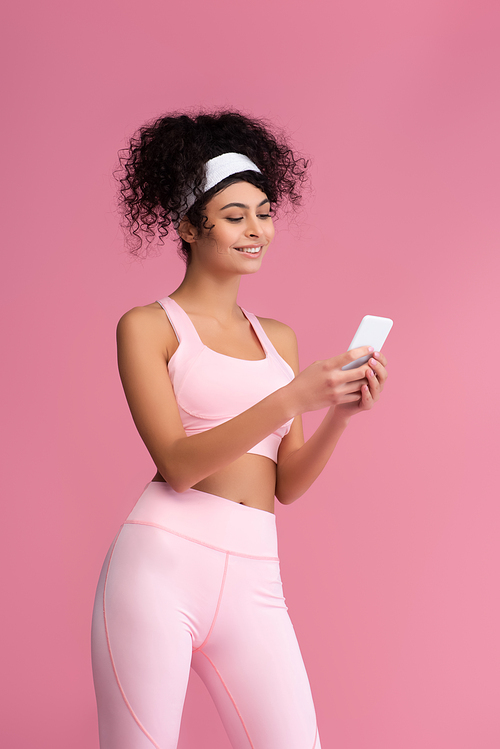 cheerful young sportswoman using smartphone isolated on pink